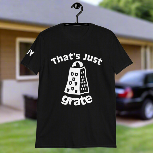 A black short-sleeve unisex t-shirt with the text 'THAT'S JUST GRATE' printed on the front, hanging on a hanger in front of a house.